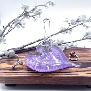 May include: A purple glass heart-shaped ring holder with a clear glass stem. The holder is sitting on a wooden surface.