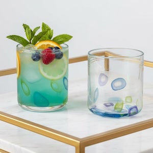May include: Two clear, round drinking glasses. One glass contains a blue-green liquid with fruit and mint. The other glass is empty, with colourful, abstract designs. Both glasses sit on a white marble tray with gold trim.