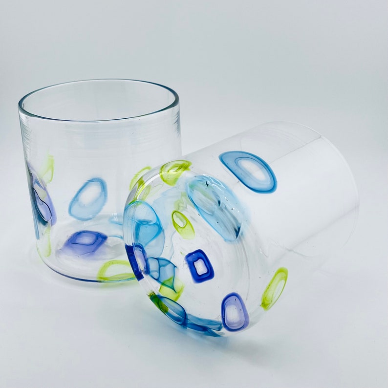 May include: Two clear glass tumblers with colourful abstract designs. One tumbler is upright, the other is on its side. The designs include blue, green, and purple shapes. The tumblers are on a white background.