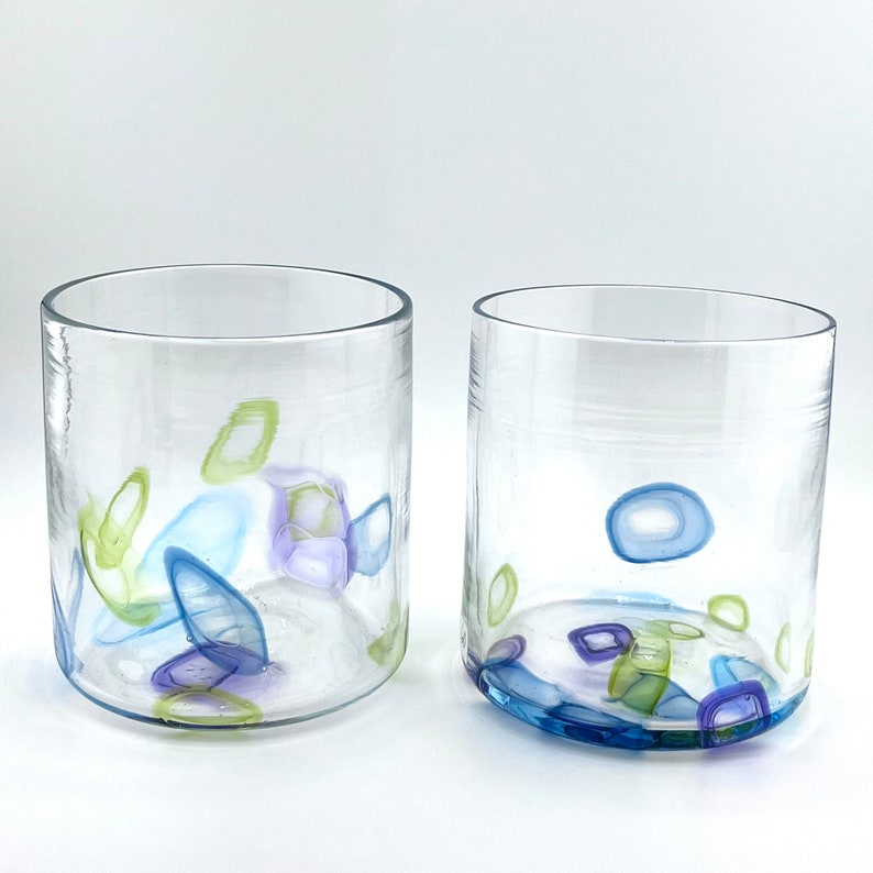 May include: Two clear glass tumblers with colourful abstract designs. The designs include blue, green, and purple shapes. The tumblers have a rounded base and straight sides, ideal for serving drinks.