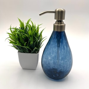 May include: A blue glass soap dispenser with a silver pump. The dispenser has a crackled finish and is shaped like a teardrop. The dispenser is sitting on a white surface next to a small potted plant.