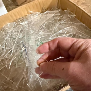 May include: A cardboard box filled with many pieces of clear, broken glass. A hand is holding a piece of the glass. The glass pieces are of various shapes and sizes. The box is sitting on a wooden surface.