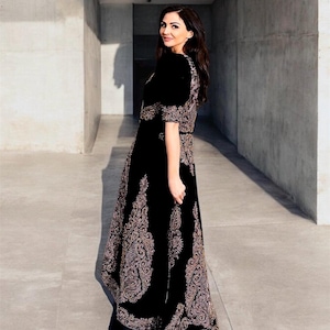 Puede incluir: Una mujer con un vestido largo de terciopelo negro con intrincados bordados dorados. El vestido tiene un corpiño ajustado y una falda fluida.