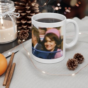 May include: White ceramic coffee mug with a photo of Jacqueline Kennedy Onassis and John F. Kennedy. The photo shows them in a car, with Jacqueline smiling and John looking forward.
