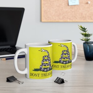 Könnte beinhalten: Zwei weiße Keramikbecher mit gelbem Hintergrund und einem schwarz-weißen Bild einer Schlange mit dem Text "Don't Tread On Me".
