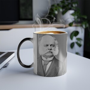 Könnte beinhalten: Eine weiße Keramiktasse mit schwarzem Henkel, auf der ein Schwarzweißbild eines Mannes mit großem Bart und Schnurrbart abgebildet ist. Die Tasse ist mit einer braunen Flüssigkeit gefüllt und steht auf einer weißen Oberfläche.