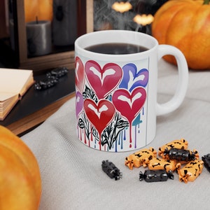 Könnte beinhalten: Weiße Keramik-Kaffeetasse mit einer bunten Grafik von tropfenden Herzen. Die Herzen sind rot, rosa, lila und blau. Die Tasse hat einen schwarzen Henkel und ist mit Kaffee gefüllt.