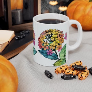 Könnte beinhalten: Eine weiße Keramiktasse mit einem bunten Blumenmuster. Die Tasse ist mit Kaffee gefüllt und Dampf steigt aus der Oberseite auf. Die Tasse steht auf einem Tisch mit einer braunen Tischdecke und etwas Süßigkeiten.