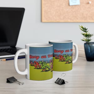 Könnte beinhalten: Zwei weiße Keramikbecher mit einer bunten Grafik von laufenden Menschen und dem Text "Keep on Truckin'" darauf gedruckt. Die Becher stehen auf einer Holzoberfläche.