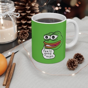 Peut inclure: Une tasse en céramique verte avec une anse blanche. La tasse présente une image de dessin animé d'une grenouille verte avec une bulle de dialogue qui dit "feels good man".
