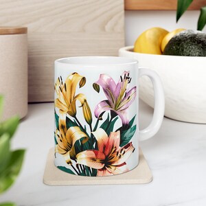Peut inclure: Une tasse en céramique blanche avec un motif floral composé de lys jaunes, roses et orange. La tasse a une grande anse et repose sur un dessous-de-tasse beige.