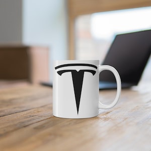 May include: White ceramic mug with a black Tesla logo printed on the front.