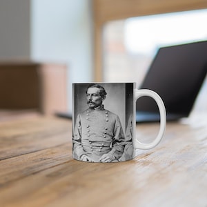 May include: A white ceramic mug with a black and white photo of a man in a military uniform. The man has a mustache and is wearing a button-up shirt with a collar. The mug has a handle on the right side.