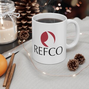 Peut inclure: Tasse en céramique blanche avec le mot "REFCO" en noir et un logo bordeaux. La tasse est remplie d'un liquide foncé et repose sur une surface gris clair. Éléments décoratifs : bougie, pommes de pin et guirlande lumineuse.