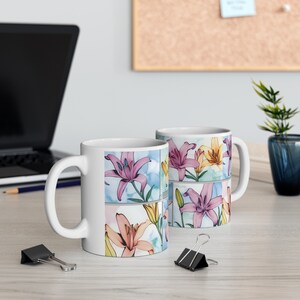 Könnte beinhalten: Zwei weiße Keramikbecher mit einem Aquarelldesign aus rosa, gelben und orangefarbenen Lilien. Die Becher haben einen Henkel auf der rechten Seite.