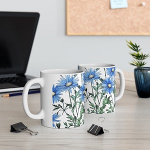 Könnte beinhalten: Zwei weiße Keramikbecher mit einem blauen Blumenmuster. Die Becher haben einen Henkel auf der rechten Seite und stehen auf einer hellbraunen Holzoberfläche.