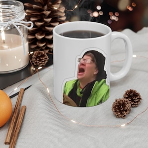 Könnte beinhalten: Weiße Keramik-Kaffeetasse mit einem bunten Bild einer Person mit Brille und grüner Jacke, die den Mund vor lauter Schreien weit aufgerissen hat.