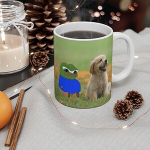 May include: White ceramic mug with a green background featuring a cartoon frog wearing a blue shirt and a golden retriever puppy. The frog and puppy are sitting in a grassy field.