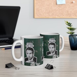 May include: Two white ceramic mugs with a green background and a black and white image of a man. The mugs have the text "I killed your nana" in a speech bubble.
