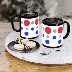Peut inclure: Deux tasses à café noires avec des pois blancs en rouge, bleu et gris. Les tasses sont remplies de café et de la vapeur s'élève du haut de chaque tasse. Les tasses sont posées sur une table en bois avec une assiette de biscuits et une cuillère.