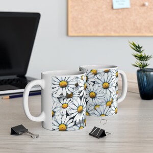 May include: Two white ceramic mugs with a daisy pattern. The mugs have a black background with white daisies and yellow centers. The mugs are on a wooden table.