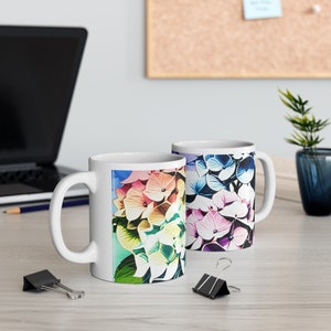 Könnte beinhalten: Zwei weiße Keramikbecher mit bunten Hortensien-Blütenmustern. Der Becher links hat eine regenbogenfarbene Hortensie, während der Becher rechts eine blaue und violette Hortensie hat.