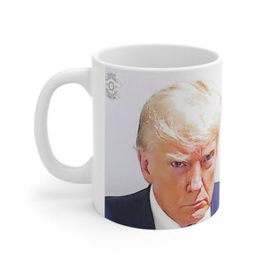 May include: White ceramic mug featuring a portrait of a man with blonde hair and a stern expression. The mug has a white handle and a printed image of a person in a suit. The mug has a Sheriff's office seal.