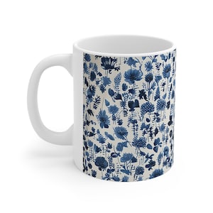 Può includere: Tazza in ceramica bianca con un motivo floreale blu e bianco. Il motivo presenta una varietà di fiori e foglie in un design ripetitivo.
