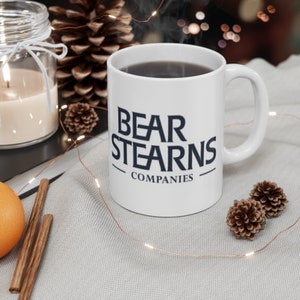 Könnte beinhalten: Eine weiße Keramiktasse mit einem schwarzen Logo, auf dem "BEAR STEARNS COMPANIES" steht. Die Tasse ist mit Kaffee gefüllt und Dampf steigt aus der Oberseite auf. Die Tasse steht auf einer weißen Oberfläche mit Lichterketten und Tannenzapfen.