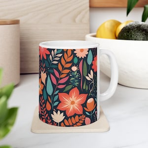 Könnte beinhalten: Weiße Keramiktasse mit einem bunten Blumenmuster. Das Muster zeigt rote, orange, gelbe, grüne und blaue Blumen und Blätter auf einem dunkelblauen Hintergrund.