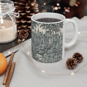 Könnte beinhalten: Weiße Keramiktasse mit einer Schwarzweißillustration einer Stadtlandschaft und Bäumen. Die Tasse ist mit Kaffee gefüllt und hat einen Henkel auf der rechten Seite.