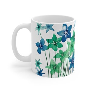 Könnte beinhalten: Weiße Keramiktasse mit einem Blumendesign aus blauen und grünen Aquarellblumen. Die Blumen sind stilisiert und haben einen verspielten Charakter.