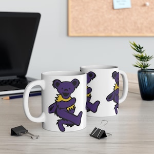 Peut inclure: Deux tasses en céramique blanche avec un motif d'ours dansant violet et jaune. L'ours porte un bandeau jaune et a un corps violet. Les tasses sont sur une table en bois.