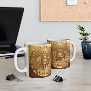 Op de afbeelding: Twee witte keramische mokken met een gouden bitcoin-ontwerp. De mokken hebben een handvat aan de rechterkant en zijn gevuld met een witte vloeistof.