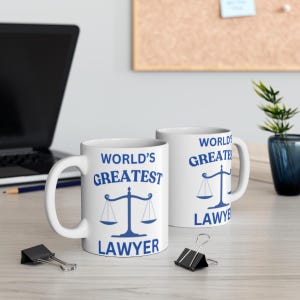 Könnte beinhalten: Zwei weiße Keramikbecher mit blauem Text und einem blauen Waagendesign. Der Text auf den Bechern lautet "World's Greatest Lawyer".