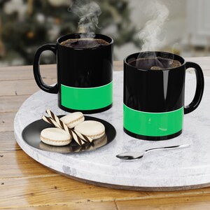 Peut inclure: Deux tasses à café noires avec des bandes vertes autour du milieu. Les tasses sont remplies de café et de la vapeur s'en échappe. Il y a trois macarons blancs et deux bâtonnets rayés noir et blanc sur une assiette noire.