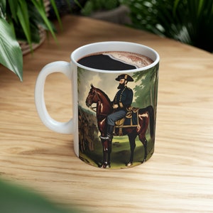 May include: White ceramic mug with a handle, featuring a vintage illustration of a man on horseback in a forest setting. The man is wearing a dark blue uniform and a black hat. The horse is brown with a white star on its forehead.