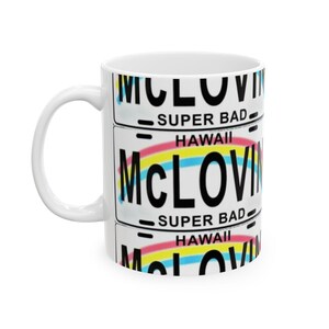 Peut inclure: Mug en céramique blanche avec un design de plaque d'immatriculation. Le design comprend le nom "McLOVIN" en noir, les mots "SUPER BAD" et "HAWAII", et un arc-en-ciel. La tasse a une anse blanche.