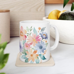 Könnte beinhalten: Weiße Keramiktasse mit einem Blumenmuster. Das Design zeigt eine Vielzahl von Blumen in den Farben Rosa, Blau, Orange und Gelb. Die Blumen sind in einem Bouquet-artigen Muster angeordnet.