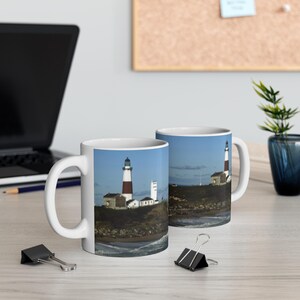 May include: Two white ceramic mugs with a lighthouse image. The mugs feature a lighthouse with a red and white striped tower on a small island with a blue sky and ocean in the background.
