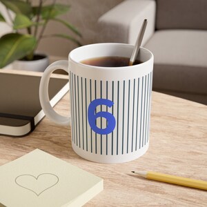Könnte beinhalten: Eine weiße Keramiktasse mit schwarzen Streifen und einer blauen Zahl 6 auf der Vorderseite. Die Tasse ist mit Kaffee gefüllt und ein silberner Löffel befindet sich in der Tasse.