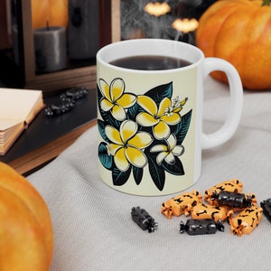 Könnte beinhalten: Weiße Keramiktasse mit einem Blumendesign mit gelben Frangipani-Blüten und grünen Blättern. Die Tasse ist mit Kaffee gefüllt und hat einen weißen Henkel.