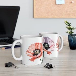 Könnte beinhalten: Zwei weiße Keramikbecher mit einem Blumendesign. Die Becher zeigen eine rosa und orangefarbene Mohnblume mit schwarzem Zentrum. Die Becher stehen auf einer weißen Oberfläche.