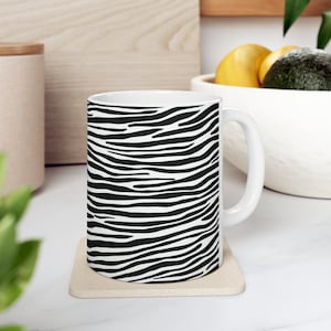 Peut inclure: Une tasse en céramique blanche avec un motif de rayures de zèbre noir et blanc. La tasse a une anse et est posée sur un dessous-de-tasse beige.