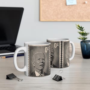 May include: Two white ceramic mugs featuring a black and white image of a person's face on a postage stamp design. The text "0c" is printed on the stamp.