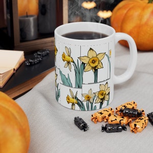 Könnte beinhalten: Weiße Keramiktasse mit einem Design aus gelben Narzissen auf weißem Hintergrund. Die Tasse ist mit Kaffee gefüllt und Dampf steigt aus der Oberseite auf.