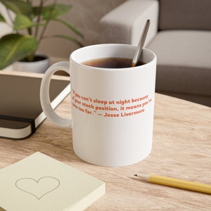 Könnte beinhalten: Eine weiße Keramik-Kaffeetasse mit schwarzem Henkel. Die Tasse hat ein Zitat in orangefarbener Schrift: "Wenn Sie nachts nicht schlafen können, weil Sie in einer schlechten Aktienposition sind, bedeutet das, dass Sie zu weit gegangen sind." - Jesse Livermore