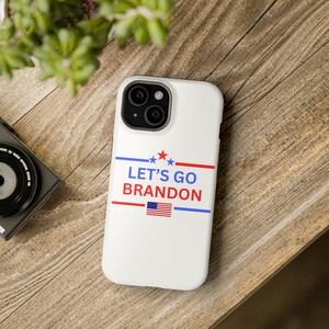 May include: White iPhone case with a red, white, and blue American flag design. The text "LET'S GO BRANDON" is printed in blue letters.
