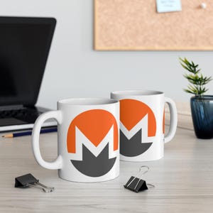 May include: Two white ceramic mugs with a black and orange logo. The logo is a stylized letter "M" inside a circle. The mugs are on a wooden surface.