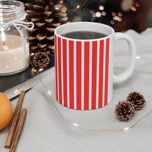Peut inclure: Une tasse en céramique blanche avec des rayures rouges et blanches. La tasse est remplie d'un liquide foncé et de la vapeur s'élève du haut. La tasse est posée sur une surface blanche avec une guirlande lumineuse.